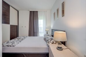 A hotel room with a bed, a nightstand with a lamp, a window with curtains, and framed pictures on the wall. - Modern 90 m2 apartment with balcony and the sea view (Split)