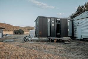 A compact, dark-colored mobile home with a large window and a door, situated in a rural setting with a bicycle and a small table nearby. - Cozy Secluded Tiny House in Warialda, New South Wales (Warialda)
