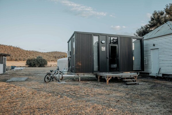 A compact, dark-colored mobile home with a large window and a door, situated in a rural setting with a bicycle and a small table nearby. - Cozy Secluded Tiny House in Warialda, New South Wales (Warialda)