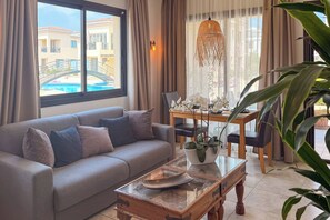 A living room with a sofa, a dining table, chairs, a hanging light fixture, and a view of a building and pool. - Sabai Pafos | Stylish Poolside Apartment with Sea View & Two Sunny Terraces (Pafos)