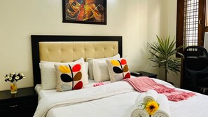 Una cama bien hecha con cabecera, varios cojines, una mesita de noche con un florero y una ventana con vistas.