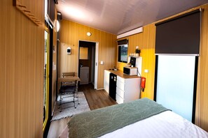 A compact room with a bed, a small kitchenette, a desk, and a mirror.