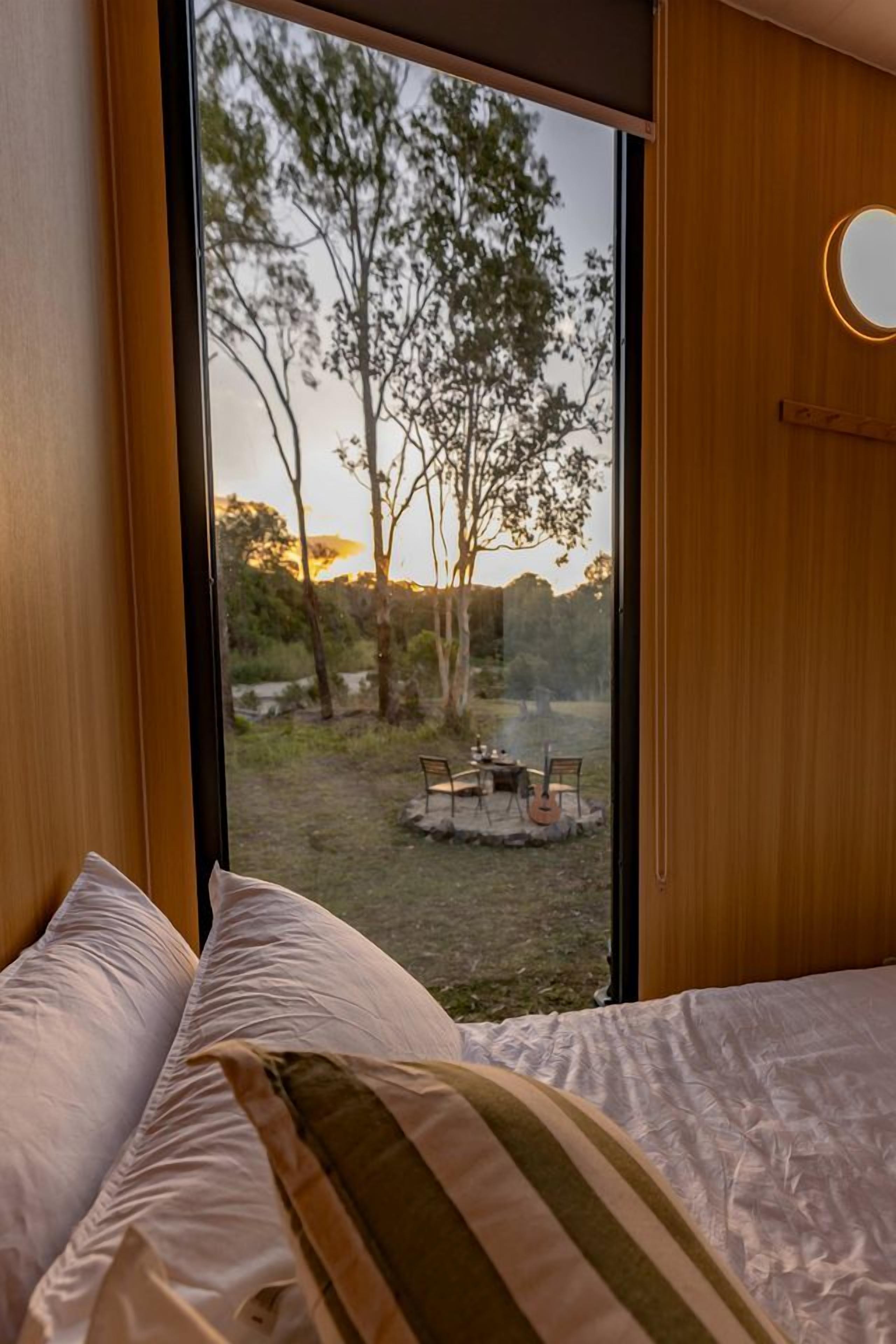 Une chambre avec une grande fenêtre, un lit avec une literie blanche et une vue sur un espace extérieur arboré, avec une table.