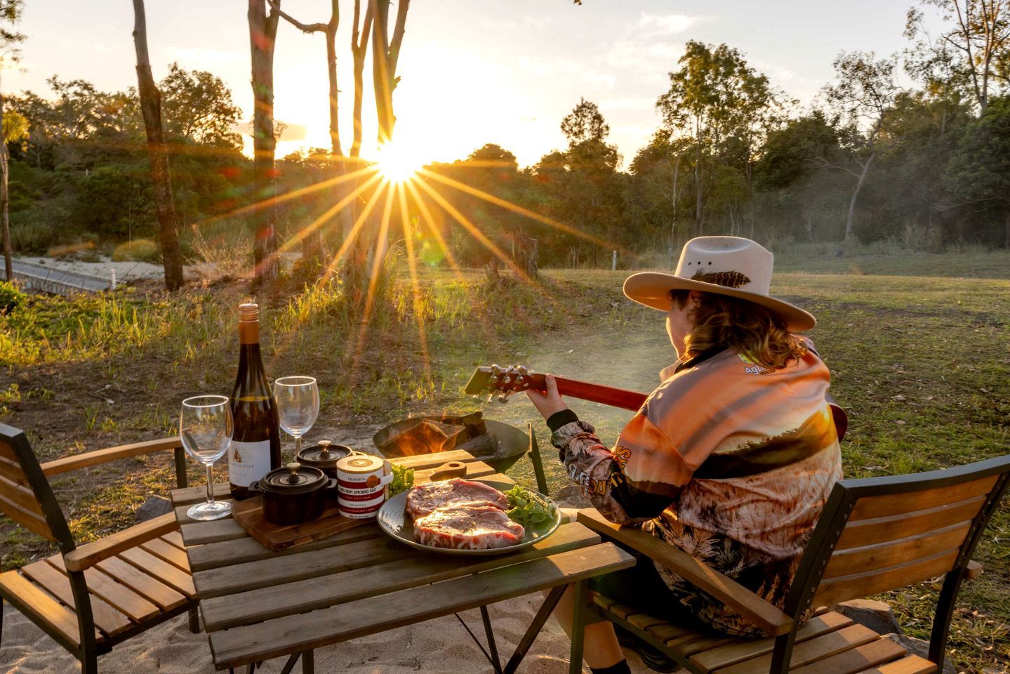 A person sitting at a wooden table outdoors with a bottle, a glass, and a plate of food.