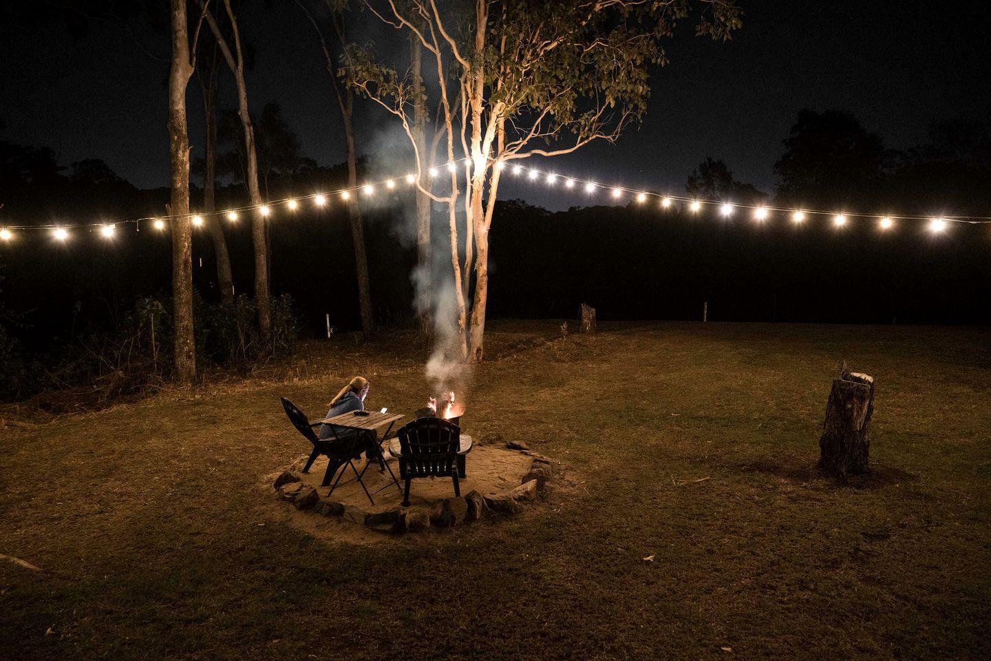 Un espace extérieur aménagé avec des sièges, un foyer et entouré d’arbres et de guirlandes lumineuses.