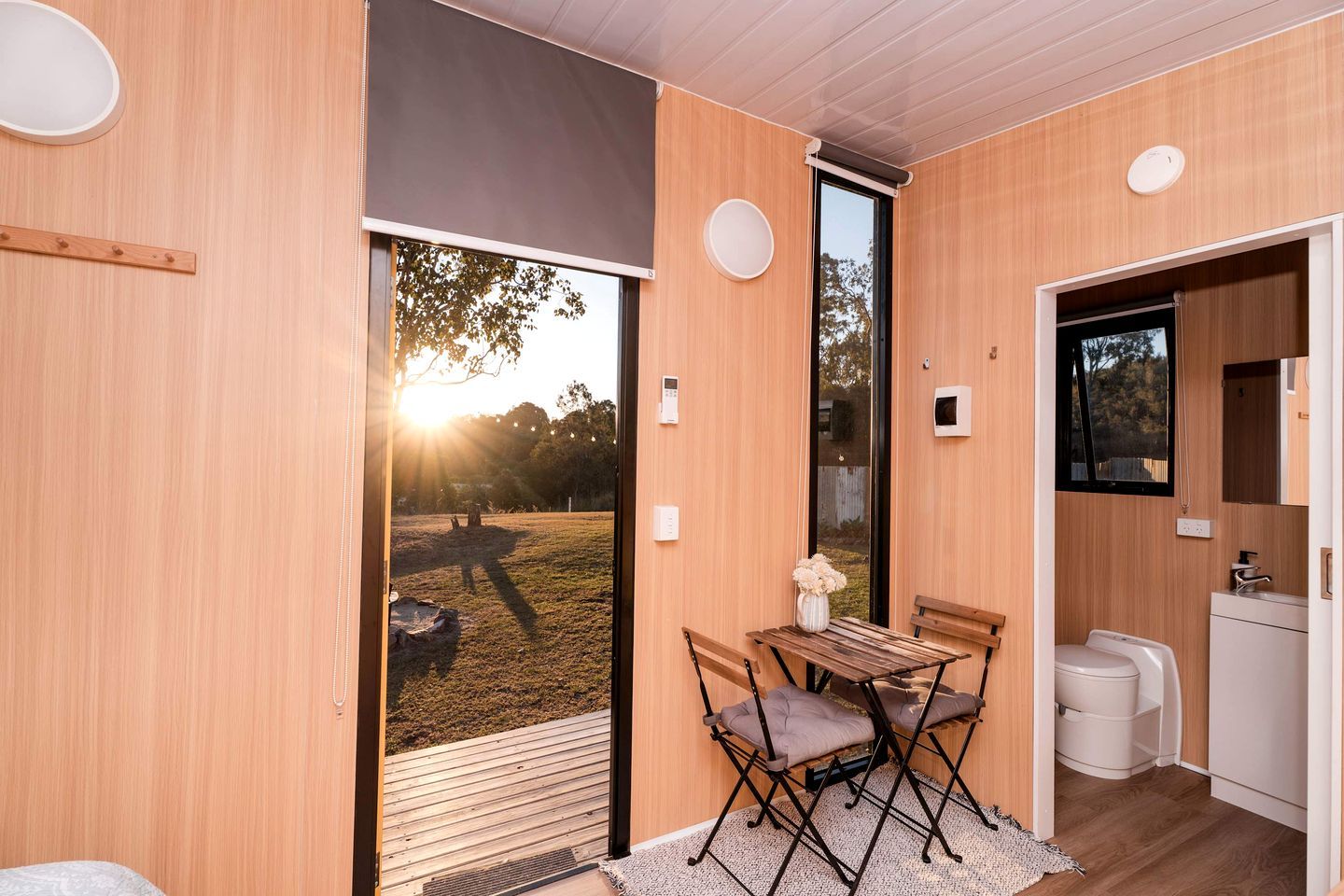 A modern interior with a wooden floor, a dining area with a table and chairs, a small kitchenette, and a sliding glass door leading to an outdoor area.