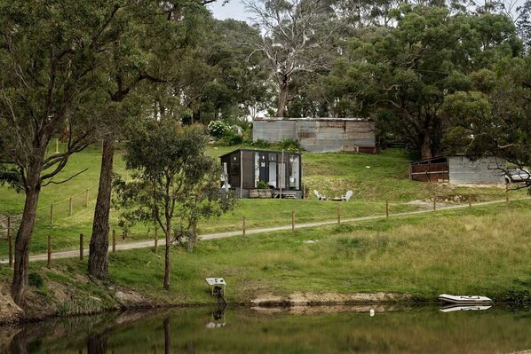 A rustic cabin with a small dock by a calm lake, surrounded by trees and a fenced yard.