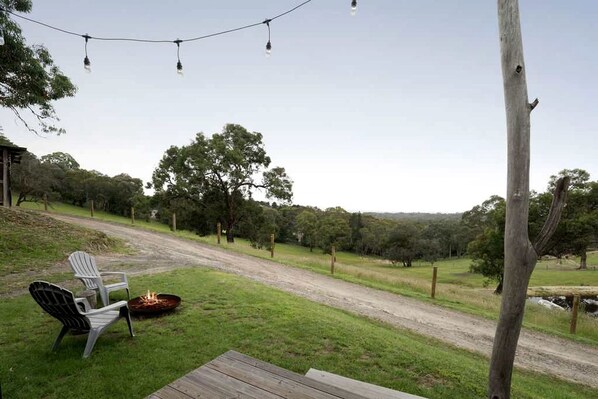 A grassy outdoor area with a wooden deck, two chairs, a fire pit, and a dirt path leading through a wooded area. - Acre Away in Hurstbridge, Victoria (Hurstbridge)