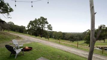 A grassy outdoor area with a wooden deck, two chairs, a fire pit, and a dirt path leading through a wooded area.