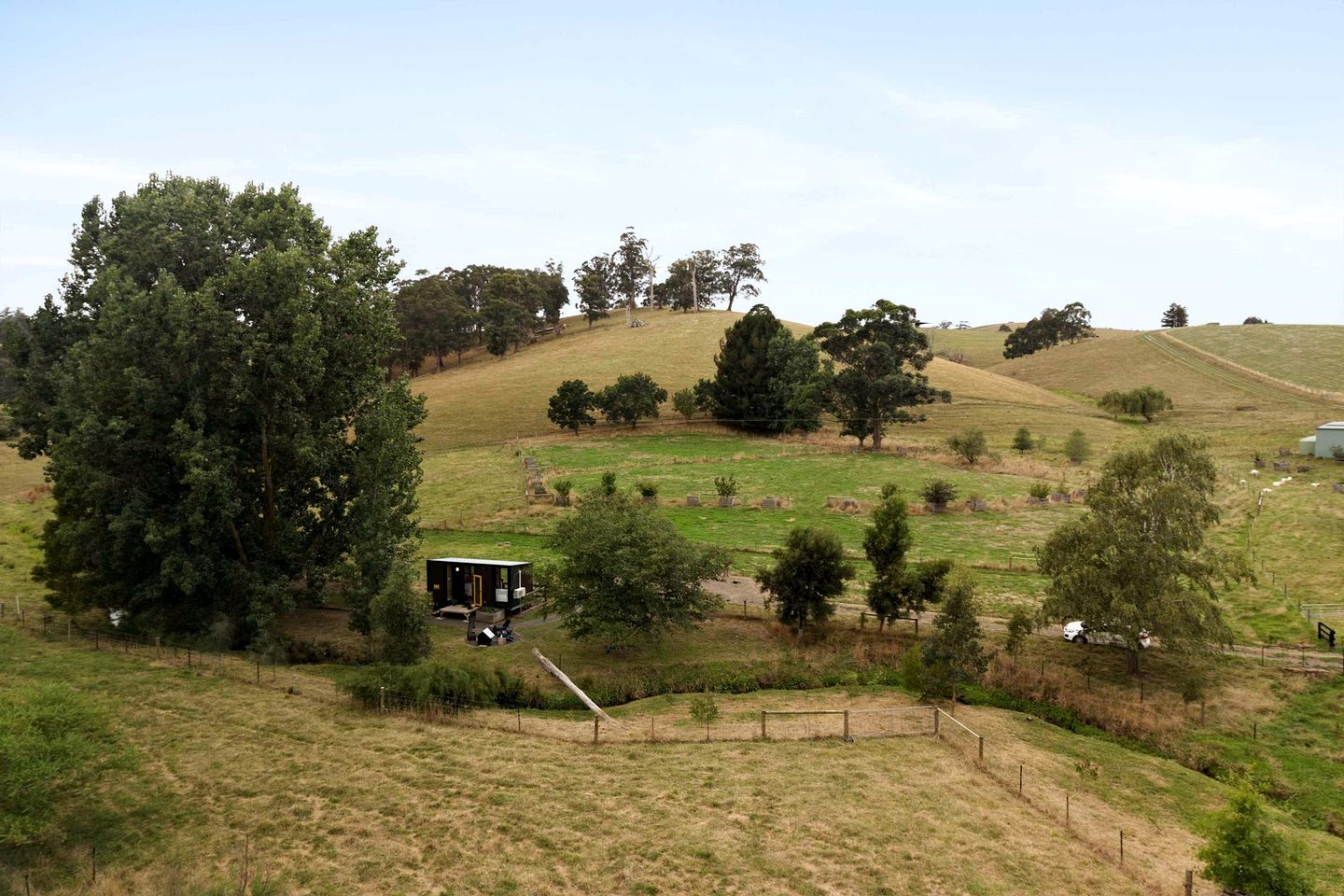 A rural landscape with rolling hills, scattered trees, and a small building with a fenced yard.