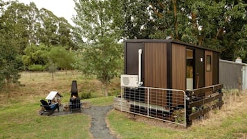 A small, single-story cabin with a metal fence, a fire pit, and a portable grill.