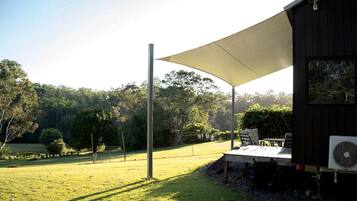 A covered outdoor seating area with a table and chairs, a fan, and a view of a grassy field and trees.