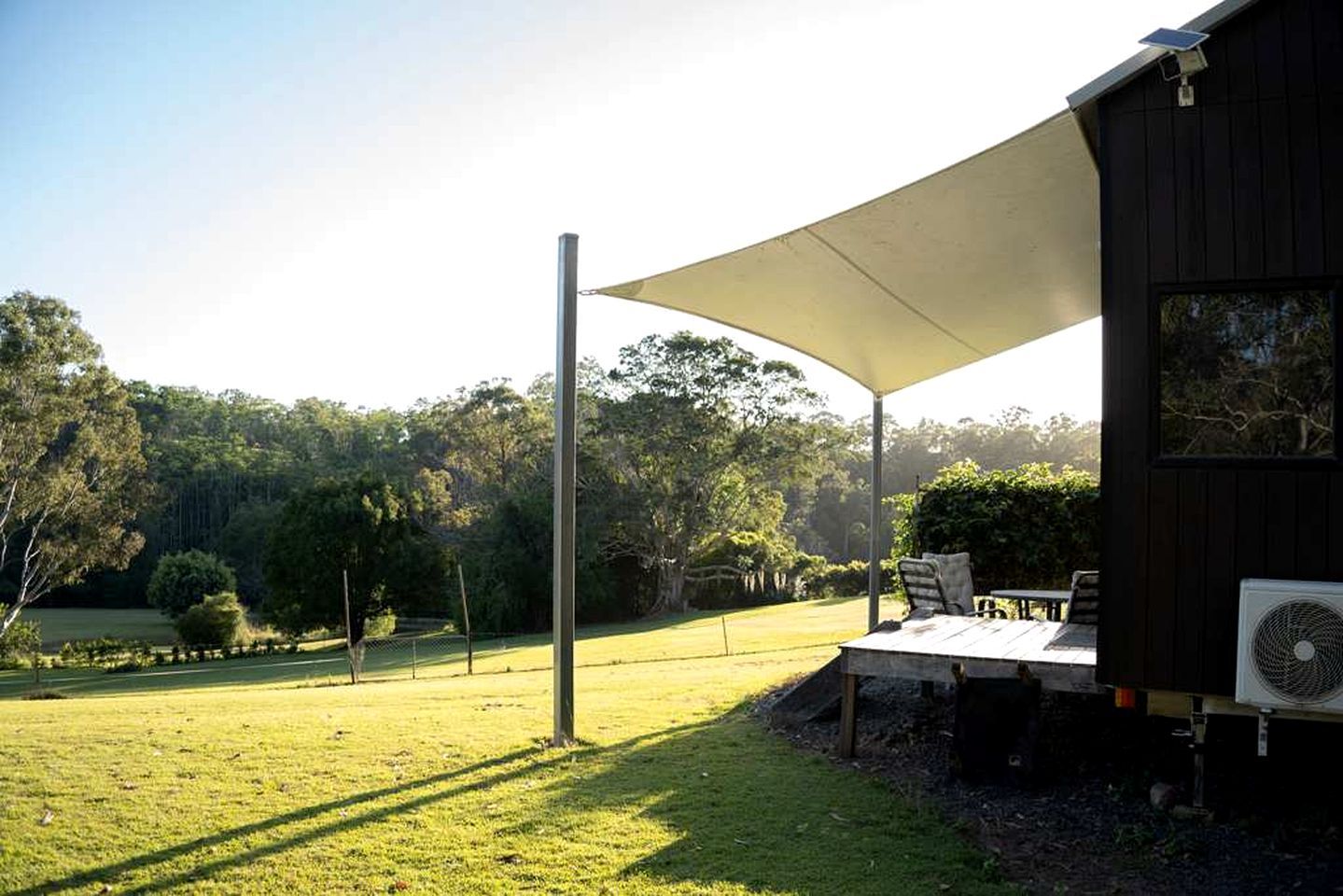 Un espacio de estar techado al aire libre con mesa y sillas, un ventilador y vista a un campo de césped y árboles.