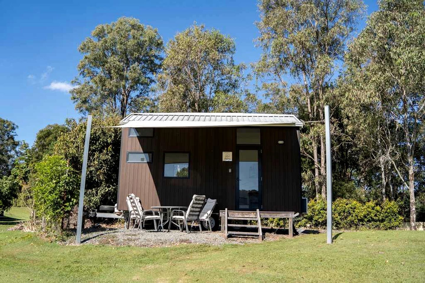 Een klein houten huisje met een metalen dak, omringd door bomen en een gazon.