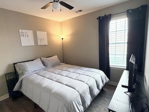Un dormitorio con cama, lámpara de pie, ventilador de techo, ventana con cortinas y dos letreros en la pared.
