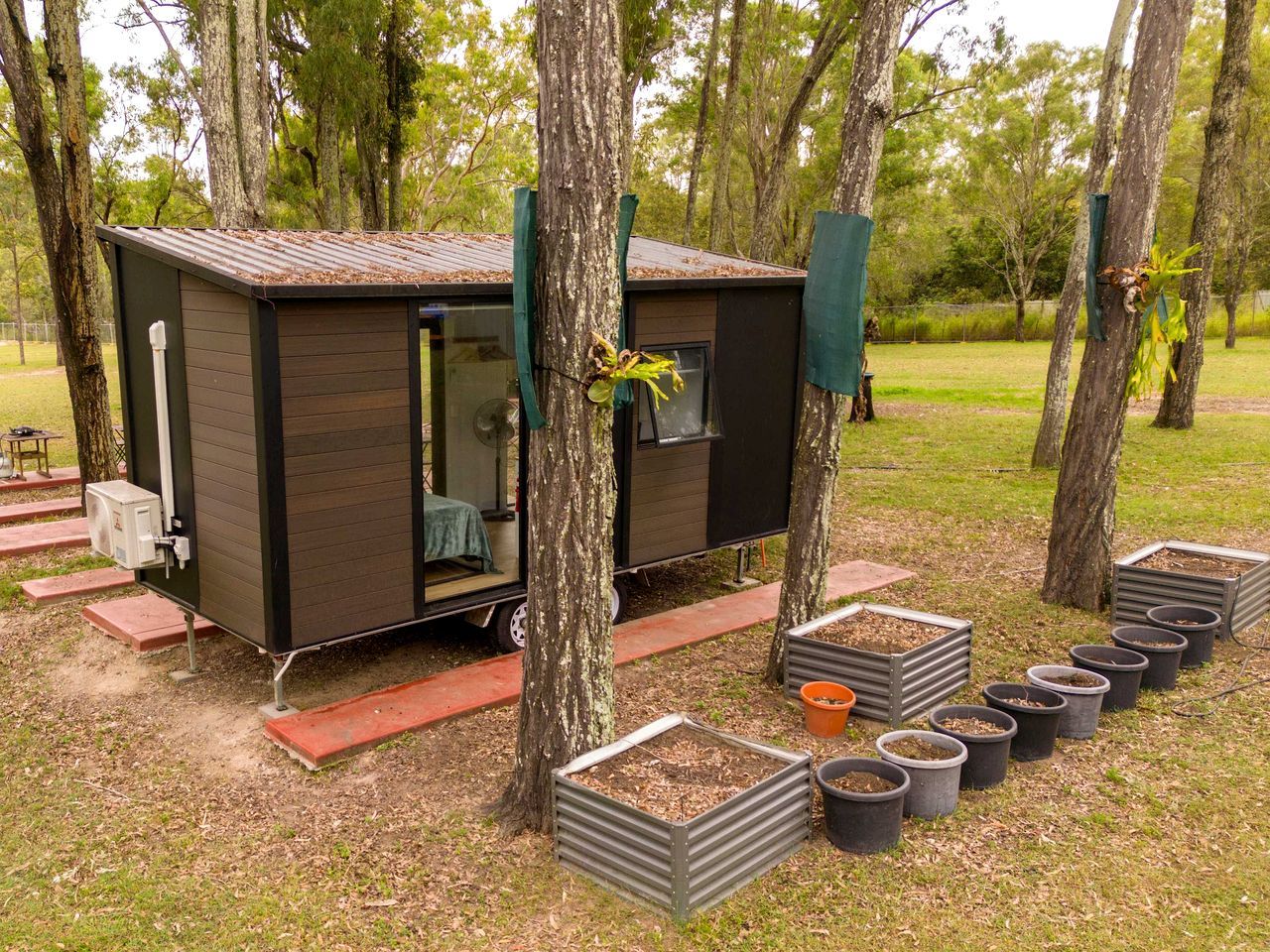 A small, dark-colored mobile home with a sliding door, surrounded by trees and potted plants.