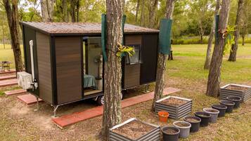 A small, dark-colored mobile home with a sliding door, surrounded by trees and potted plants.