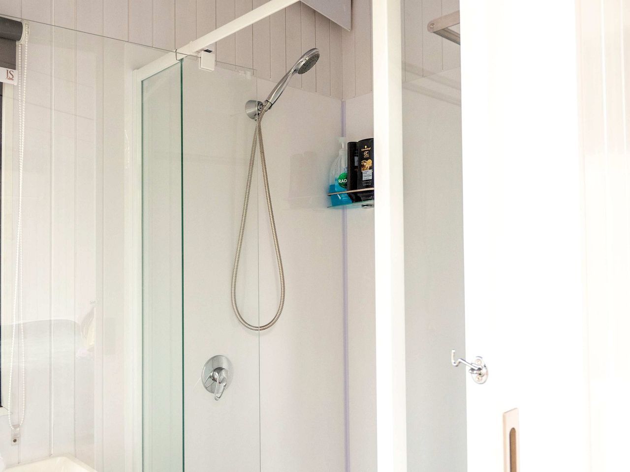 A glass-enclosed shower with a handheld showerhead, a glass door, and a small shelf with toiletries.