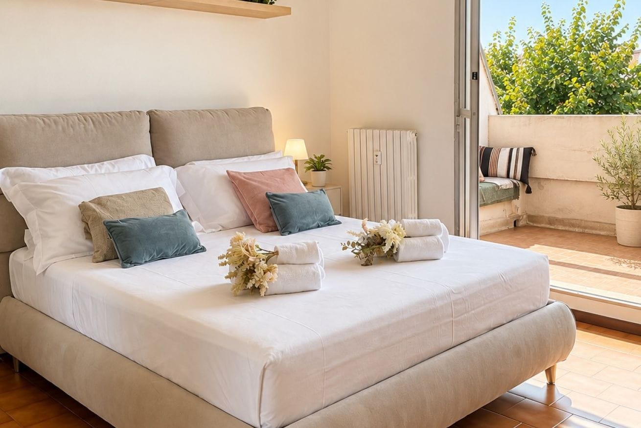 A neatly made bed with pillows and towels, a bedside lamp, and a view of a balcony with a plant.