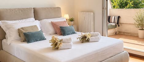 Una cama bien hecha con almohadas y toallas, una lámpara de noche y vistas a un balcón con una planta.