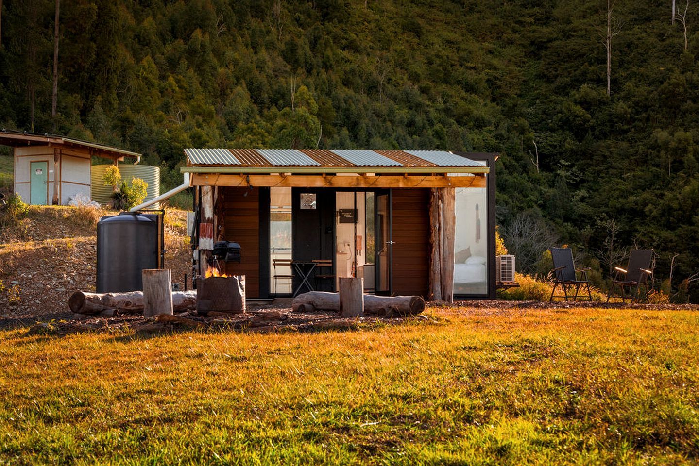 A rustic cabin with a wooden exterior and a metal roof, situated in a grassy field with a forested hill in the background.