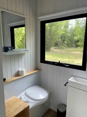 A compact bathroom with a toilet, a small shelf, a window, and a trash bin.