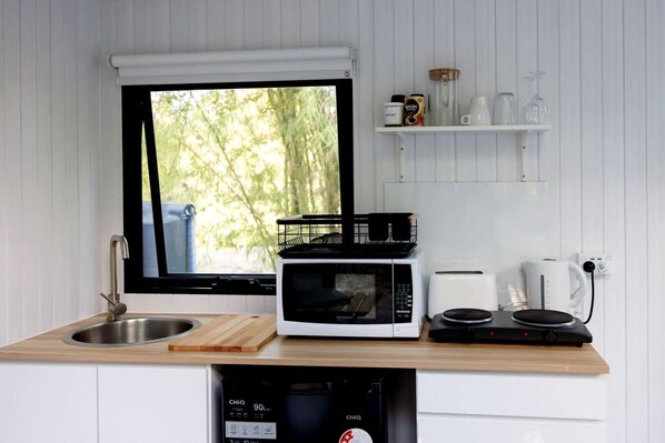 A compact kitchen with a sink, a microwave, a toaster, a kettle, and a window with a view of greenery.