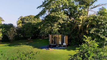 Une petite construction indépendante, dotée d’une porte et de fenêtres, entourée d’une végétation luxuriante et d’arbres.