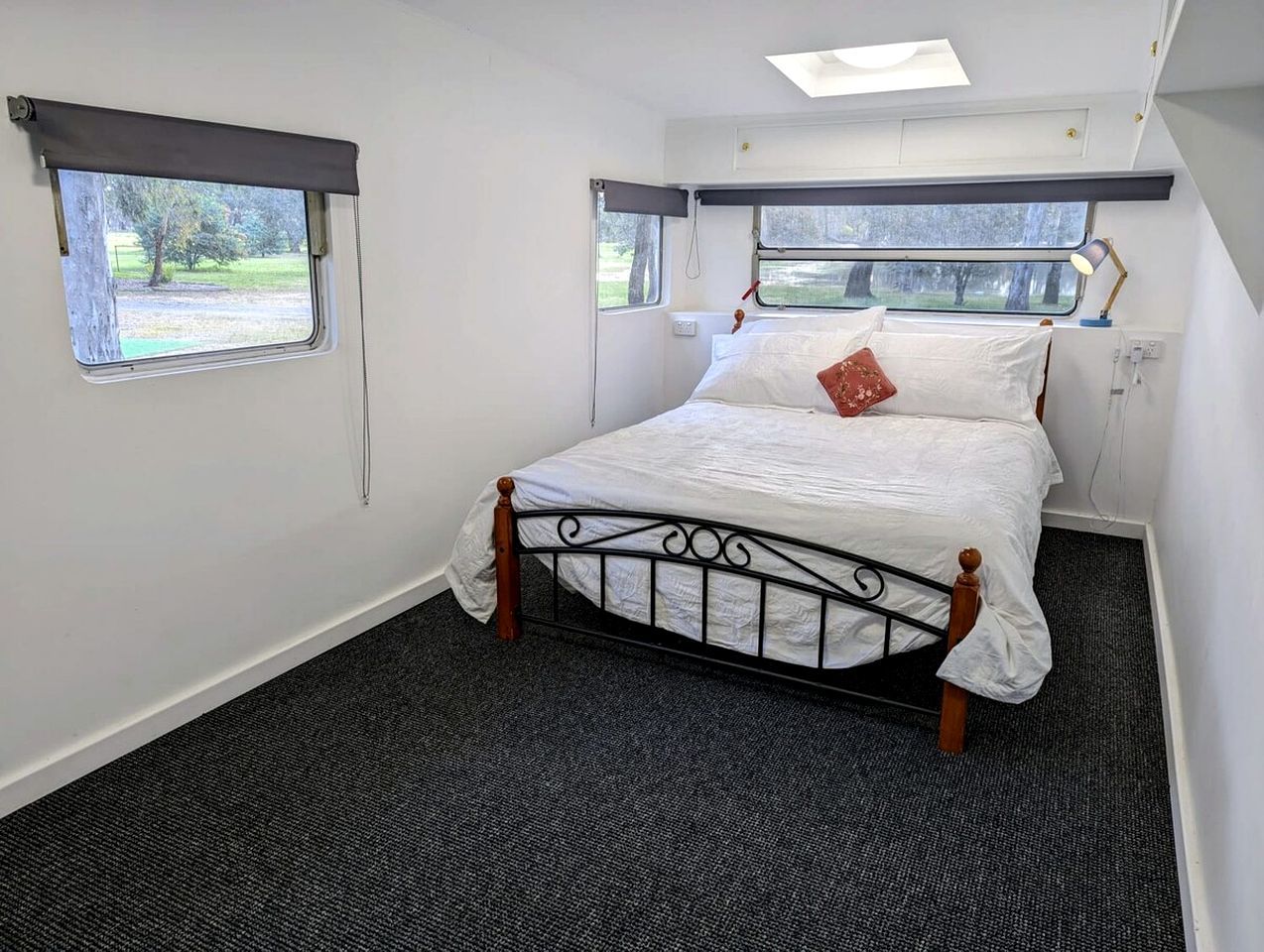 A single bed with a metal frame and wooden headboard in a room with carpeted flooring and two windows with curtains.