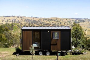 A small, dark-colored mobile home with a single window and a door, situated on a trailer with a grassy field and hills in the background.