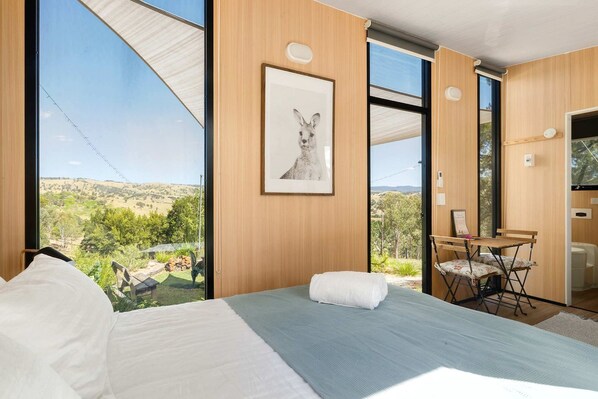 A bedroom with a large window, a bed, a framed picture, and a small table with chairs.