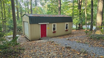 Une petite cabane en bois avec une porte rouge, une fenêtre agrémentée d’une jardinière et un chemin de gravier menant à l’entrée.