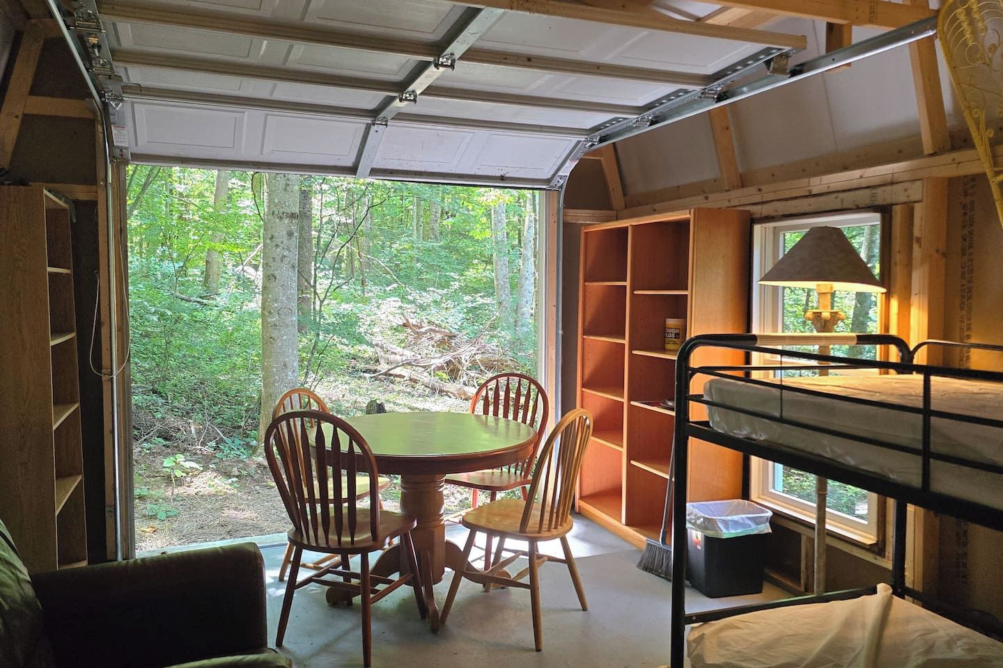 A compact room with a dining table and chairs, a bunk bed, and a window overlooking a forest.