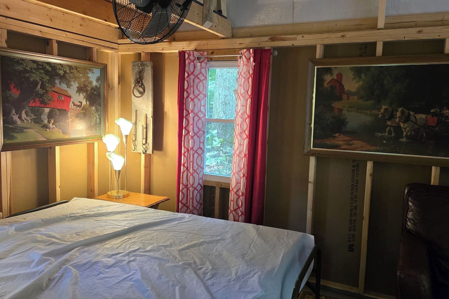 A room with a bed, a desk with a lamp, a window with curtains, and two paintings on the walls.