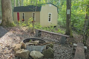 A small cabin with a fire pit and rocks in front, surrounded by trees.