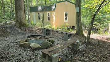 A rustic cabin with a fire pit and benches in a wooded area.