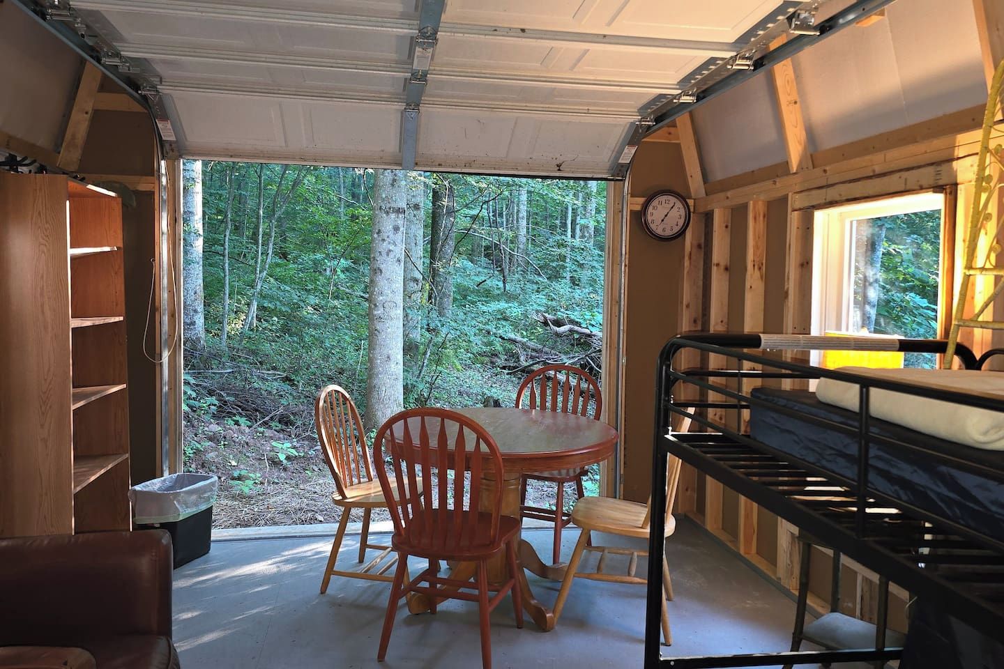 A compact, wood-paneled interior with a dining table and chairs, a bunk bed, and a view of a forested area through a large window.