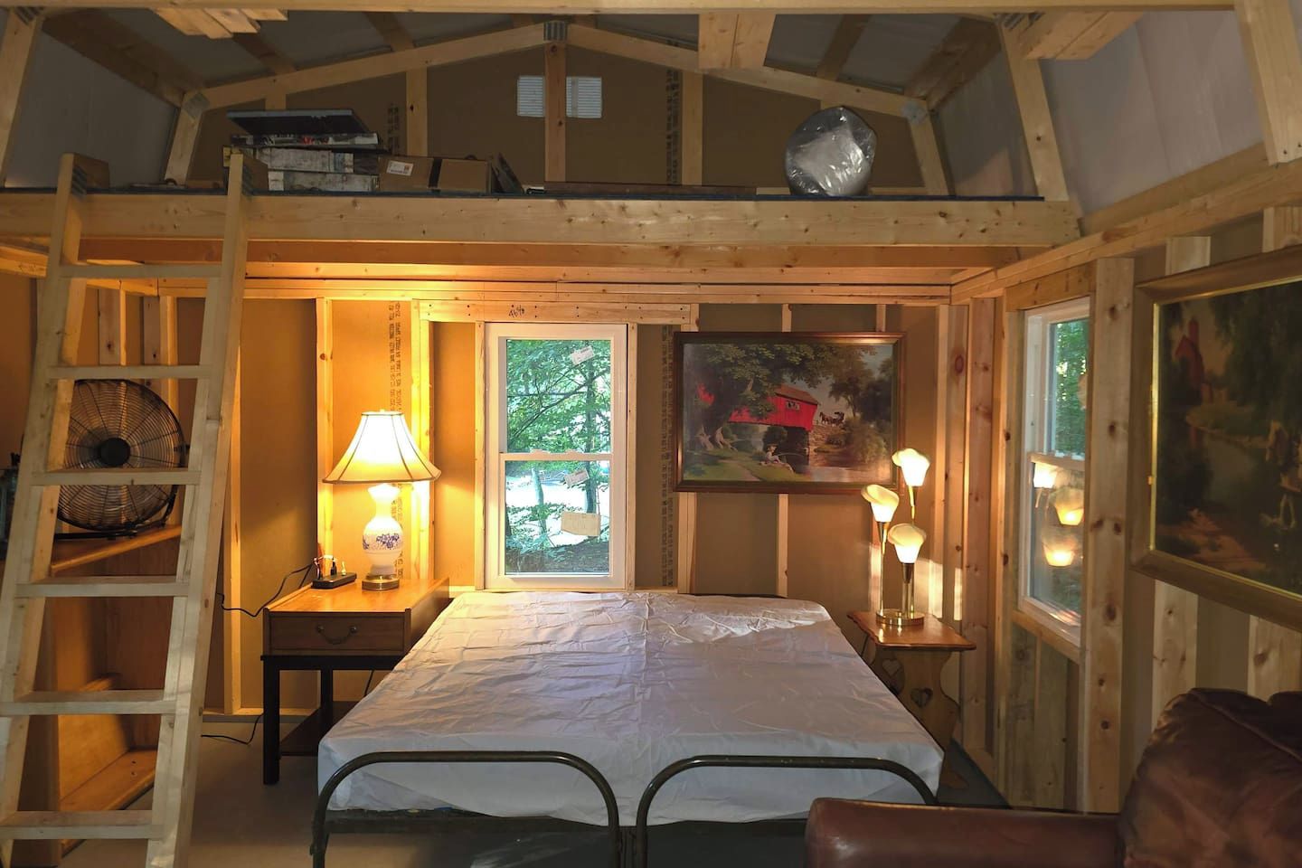 A cozy wooden cabin room with a loft bed, a small table with a lamp, a ladder, and a window with a view of greenery.