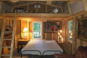 A cozy wooden cabin room with a loft bed, a small table with a lamp, a ladder, and a window with a view of greenery.