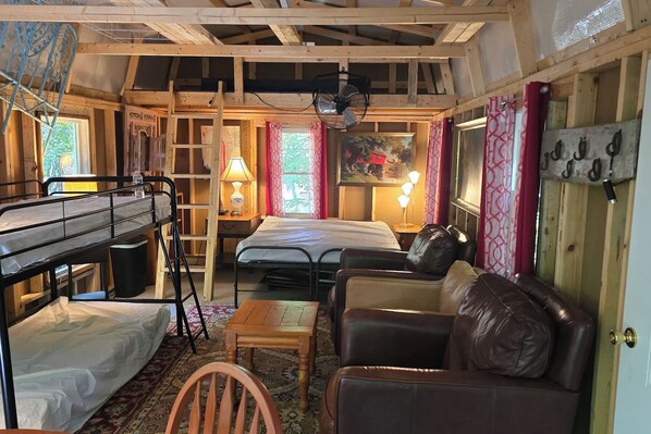Interior de una cabaña con literas, una cama individual, un sofá de cuero, una mesita, una silla, una escalera, un ventilador y una ventana con cortinas.