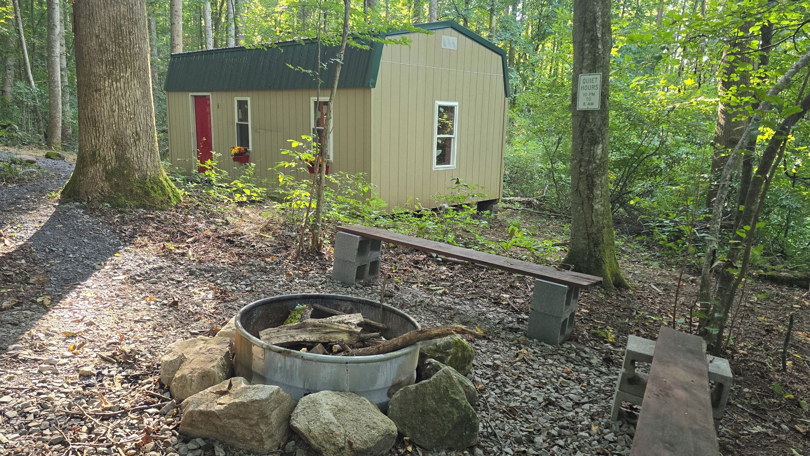 A small cabin with a red door, surrounded by trees and a fire pit with rocks.