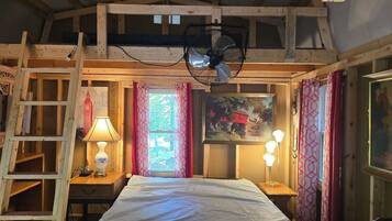 A room with a bed, a fan, a lamp, a ladder, a window with curtains, and a painting on the wall.