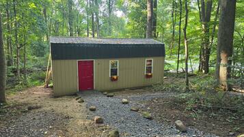 A small wooden shed with a red door, two windows, and flower boxes, surrounded by a forest.