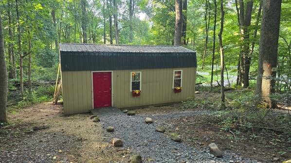 A small wooden shed with a red door, two windows, and flower boxes, surrounded by a forest.