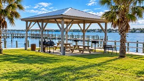 A gazebo with picnic tables and a grill, overlooking a body of water with docks and houses in the background. - Barefoot Hideaway | Beach + Pool + Boat Friendly + Lagoon | Sea Oats (Gulf Shores)