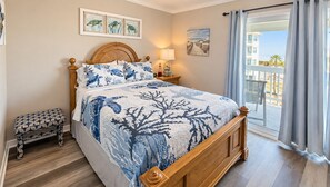 A bedroom with a bed, a bedside table, a bench, a window with curtains, and artwork on the walls. - Barefoot Hideaway | Beach + Pool + Boat Friendly + Lagoon | Sea Oats (Gulf Shores)