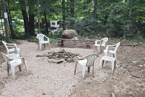 A campsite with a fire pit surrounded by plastic chairs, a large rock, and a white RV in a wooded area. - D1 Dryfork Forest Retreat-electric-heat (Parsons)