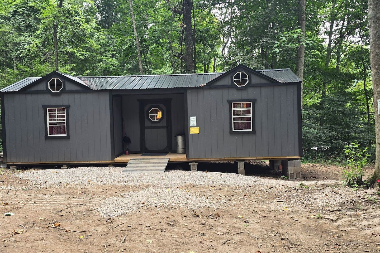 Une petite cabane de couleur sombre, dotée d’une terrasse en bois, d’une porte d’entrée et de deux fenêtres.