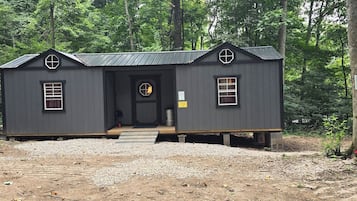 Une petite cabane de couleur sombre, dotée d’une terrasse en bois, d’une porte d’entrée et de deux fenêtres.