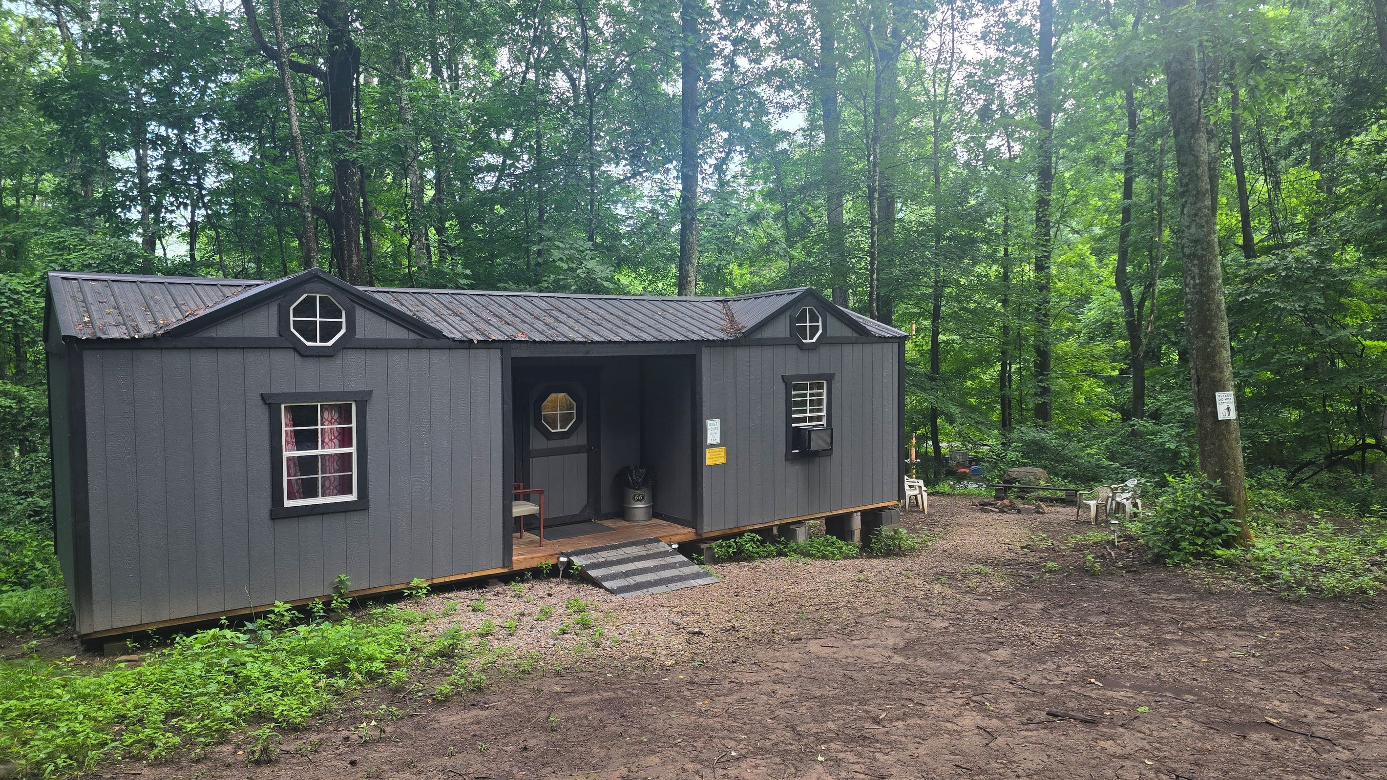 A small, dark-colored cabin with a single door and two windows, surrounded by a forest.